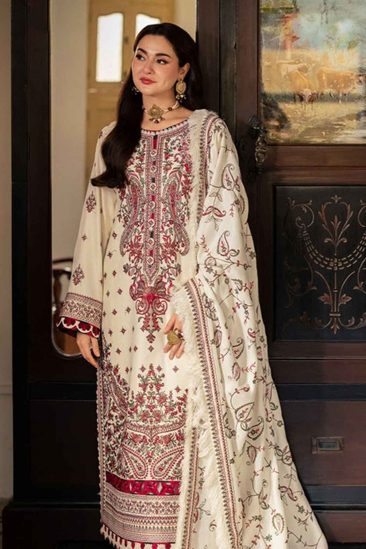 A woman models a traditional three-piece salwar kameez suit in cream with intricate red and gold embroidery, standing in an elegant indoor setting with dark wood paneling.
