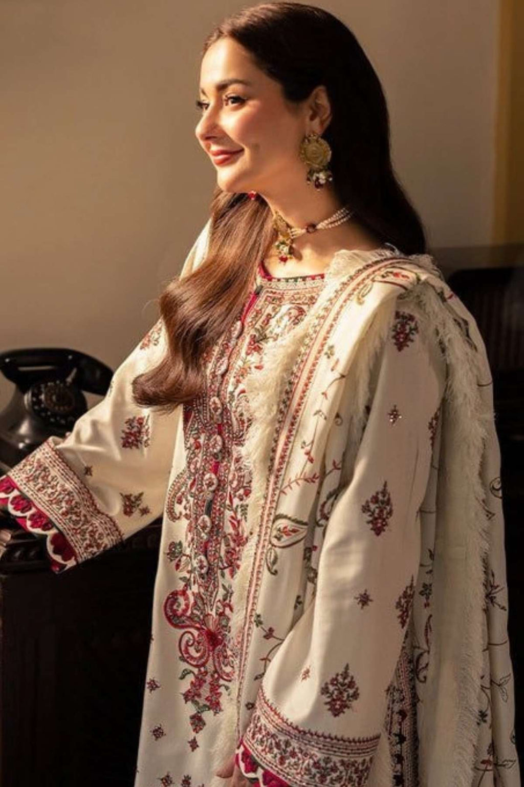 A woman wearing a traditional embroidered outfit in cream with red and green patterns, styled with ornate gold jewelry in an indoor setting.
