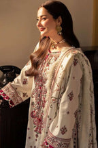 A woman wearing a traditional embroidered outfit in cream with red and green patterns, styled with ornate gold jewelry in an indoor setting.