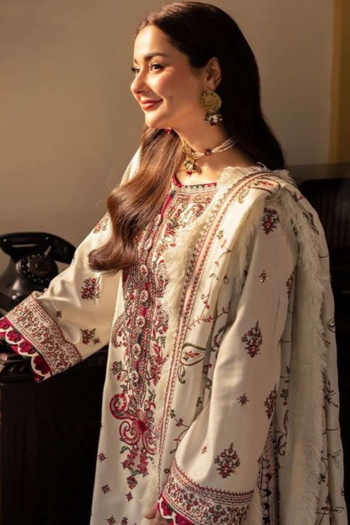 A woman wearing a traditional embroidered outfit in cream with red and green patterns, styled with ornate gold jewelry in an indoor setting.
