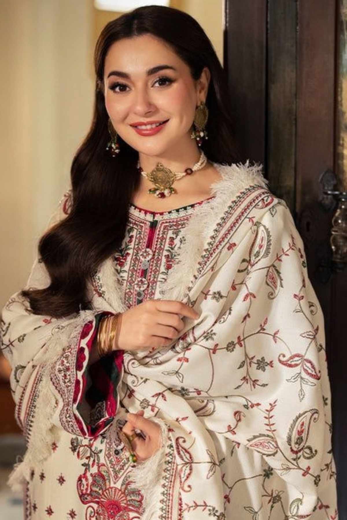 A woman wearing a cream-colored embroidered traditional outfit with red and green paisley patterns, a matching embroidered dupatta, and gold jewelry, posed in front of a dark wooden door.