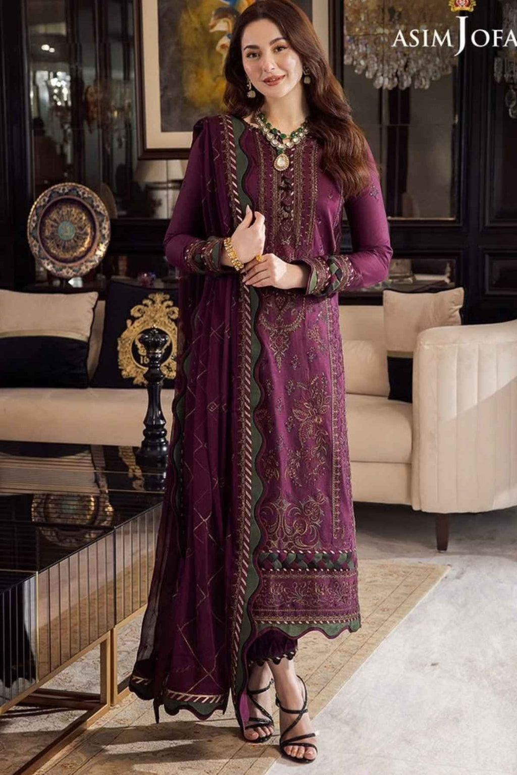 A woman models a traditional three-piece ethnic suit in a luxurious living room setting. The suit is a deep plum purple with intricate gold and green embroidery, featuring a long kurta, matching trousers, and a sheer dupatta.