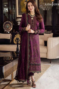 A woman models a traditional three-piece ethnic suit in a luxurious living room setting. The suit is a deep plum purple with intricate gold and green embroidery, featuring a long kurta, matching trousers, and a sheer dupatta.
