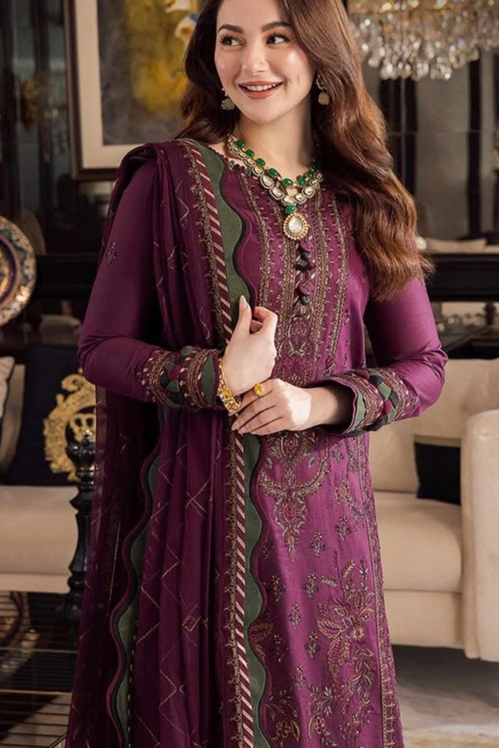A woman models a traditional Indian salwar kameez suit in deep purple with green and gold embroidery, featuring a matching dupatta, set against an elegant indoor background.