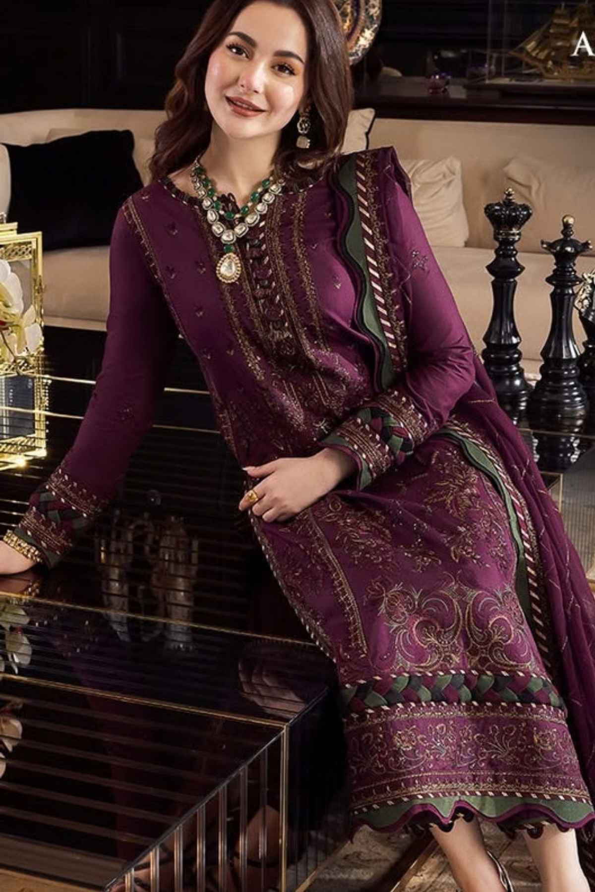 A woman models a deep purple ethnic three-piece suit with intricate gold embroidery and green accents, posing in a luxurious indoor setting.