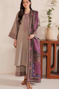 A woman models a traditional three-piece salwar kameez suit in taupe beige, featuring a matching purple embroidered dupatta draped over her shoulder, set against a warm indoor background with a wooden side table and potted plants.