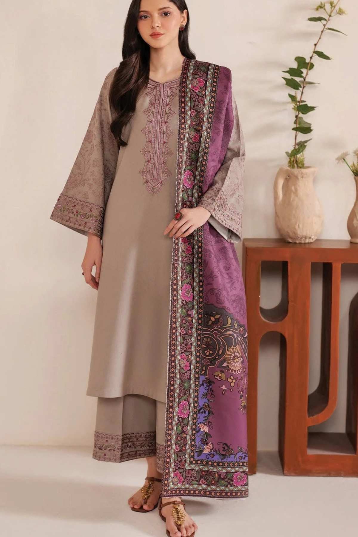 A woman models a traditional three-piece salwar kameez suit in taupe beige, featuring a matching purple embroidered dupatta draped over her shoulder, set against a warm indoor background with a wooden side table and potted plants.