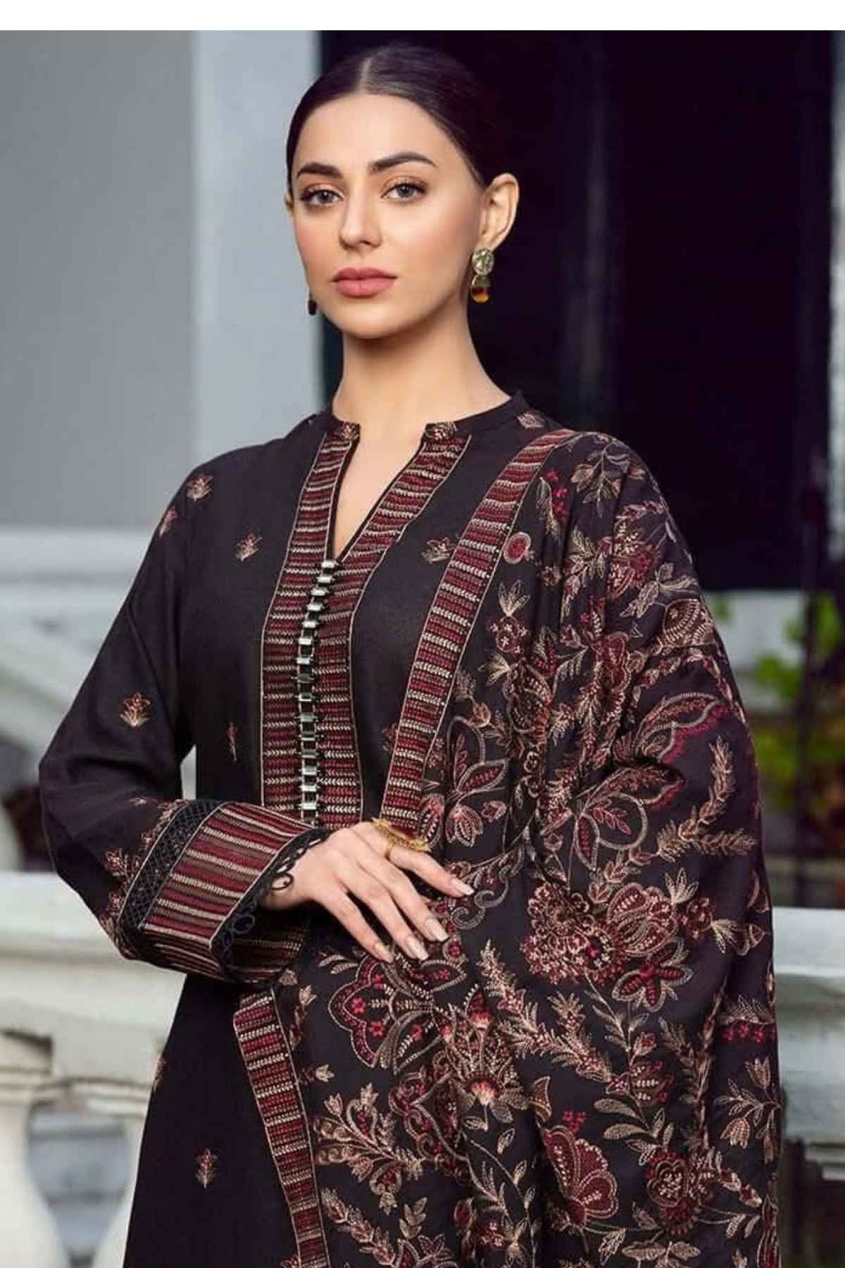 A woman modeling a black three-piece salwar kameez suit with intricate red and beige floral embroidery and striped border details, set against a blurred outdoor background.