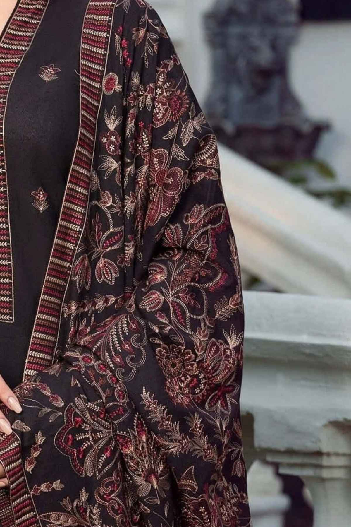 A close-up of a dark charcoal black fabric garment with intricate red, brown, and beige floral embroidery and a striped border, worn by a model in an outdoor setting.