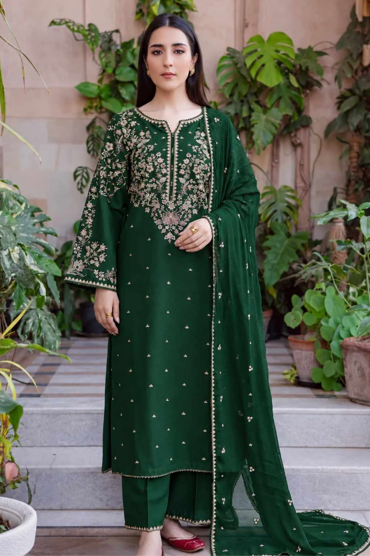 A woman models a dark green ethnic salwar kameez suit with gold embroidery, matching trousers, and a dupatta, set against a background of potted plants and stone steps.