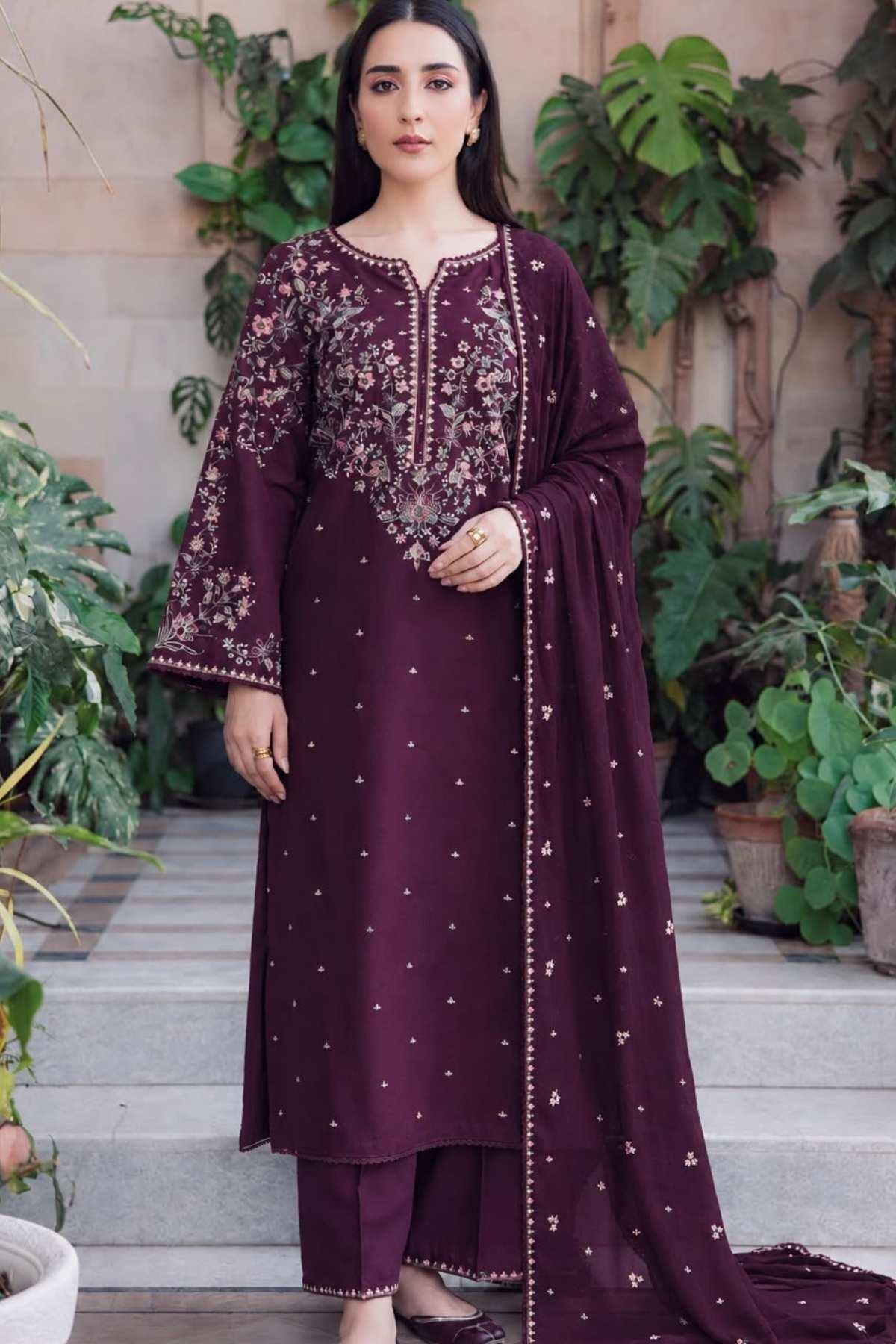 A woman models a deep purple three-piece salwar kameez suit with intricate gold embroidery on the chest and sleeves, paired with matching embroidered dupatta and trousers, set against a background of green plants and stone steps.