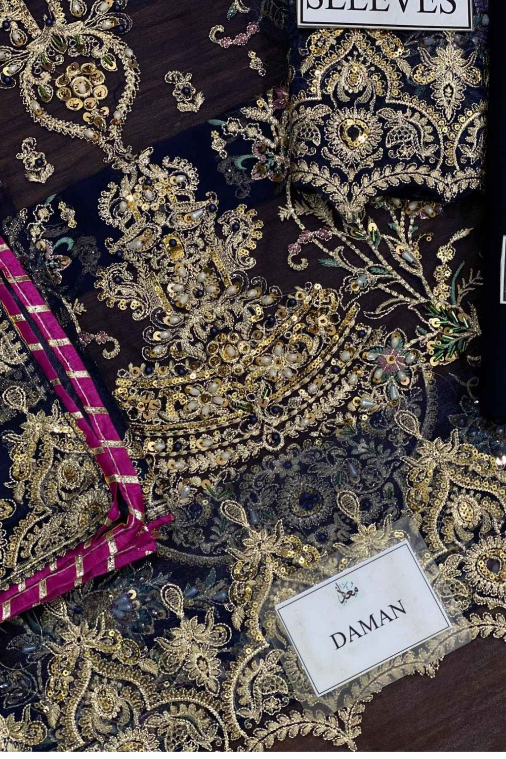 Close-up of an ornate embroidered fabric with intricate gold and beige paisley and floral patterns on a dark base, plus a magenta striped fabric strip and labels reading 'SEEVES' and 'DAMAN'.