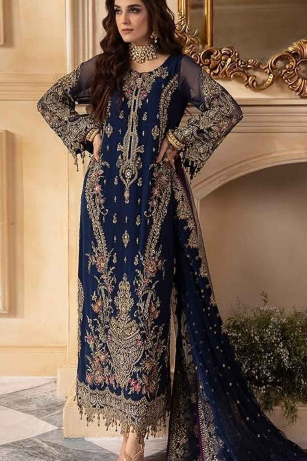 A woman models a navy blue traditional ethnic suit with intricate gold embroidery and floral patterns, featuring sheer long sleeves and a matching dupatta, posed in front of an ornate beige wall and mirror.