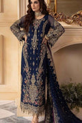 A woman models a navy blue traditional ethnic suit with intricate gold embroidery and floral patterns, featuring sheer long sleeves and a matching dupatta, posed in front of an ornate beige wall and mirror.