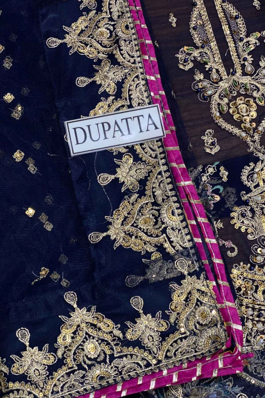 A close-up of a navy blue dupatta featuring intricate gold zari embroidery and a vibrant magenta border, with a white label reading 'DUPATTA' stitched on it.