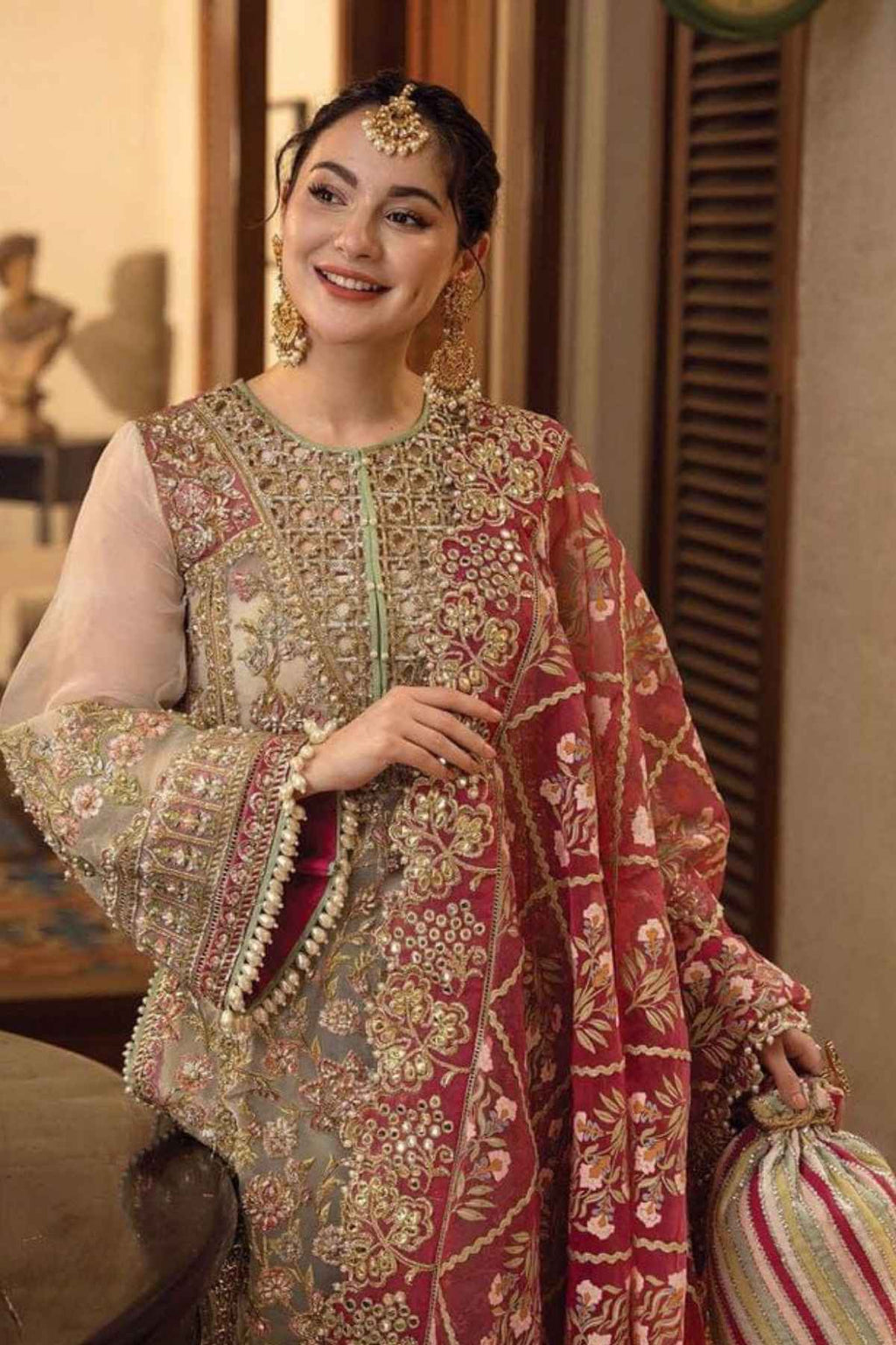 A woman wearing an ornate traditional South Asian outfit with heavy gold embroidery, a red and gold patterned dupatta, and a beige embroidered tunic, posed in an indoor setting.