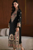 A woman models a black long kurta with elaborate gold, pink, and red embroidery, standing in front of a dark wood-paneled wall and a light gray sofa.
