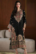 A woman models a black three-piece traditional South Asian salwar suit with elaborate gold, pink, and red embroidery. The set includes a long kurta with a high neckline, wide sleeves, and a heavily embellished hem, paired with matching trousers and a folded dupatta, set against a warm brown wooden wall and a gray sofa.