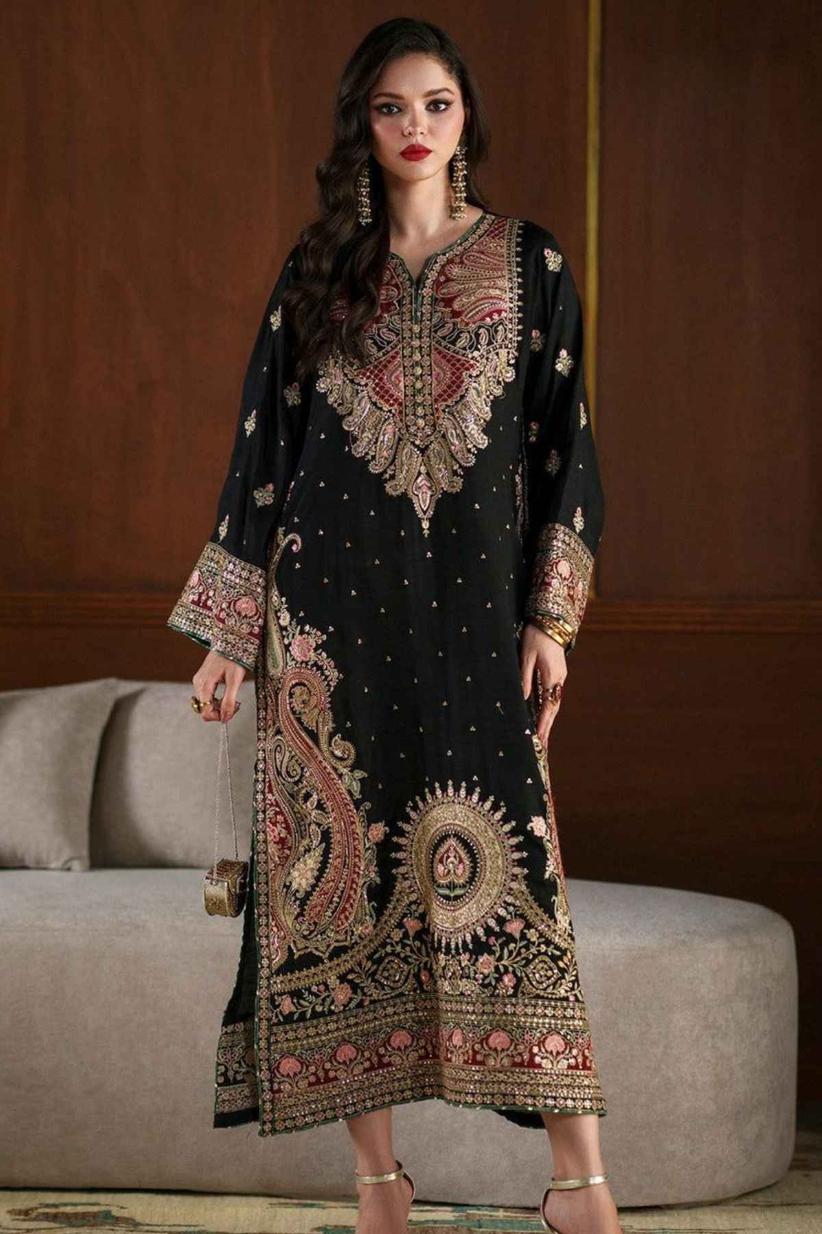 A woman models a black three-piece traditional South Asian salwar suit with elaborate gold, pink, and red embroidery. The set includes a long kurta with a high neckline, wide sleeves, and a heavily embellished hem, paired with matching trousers and a folded dupatta, set against a warm brown wooden wall and a gray sofa.
