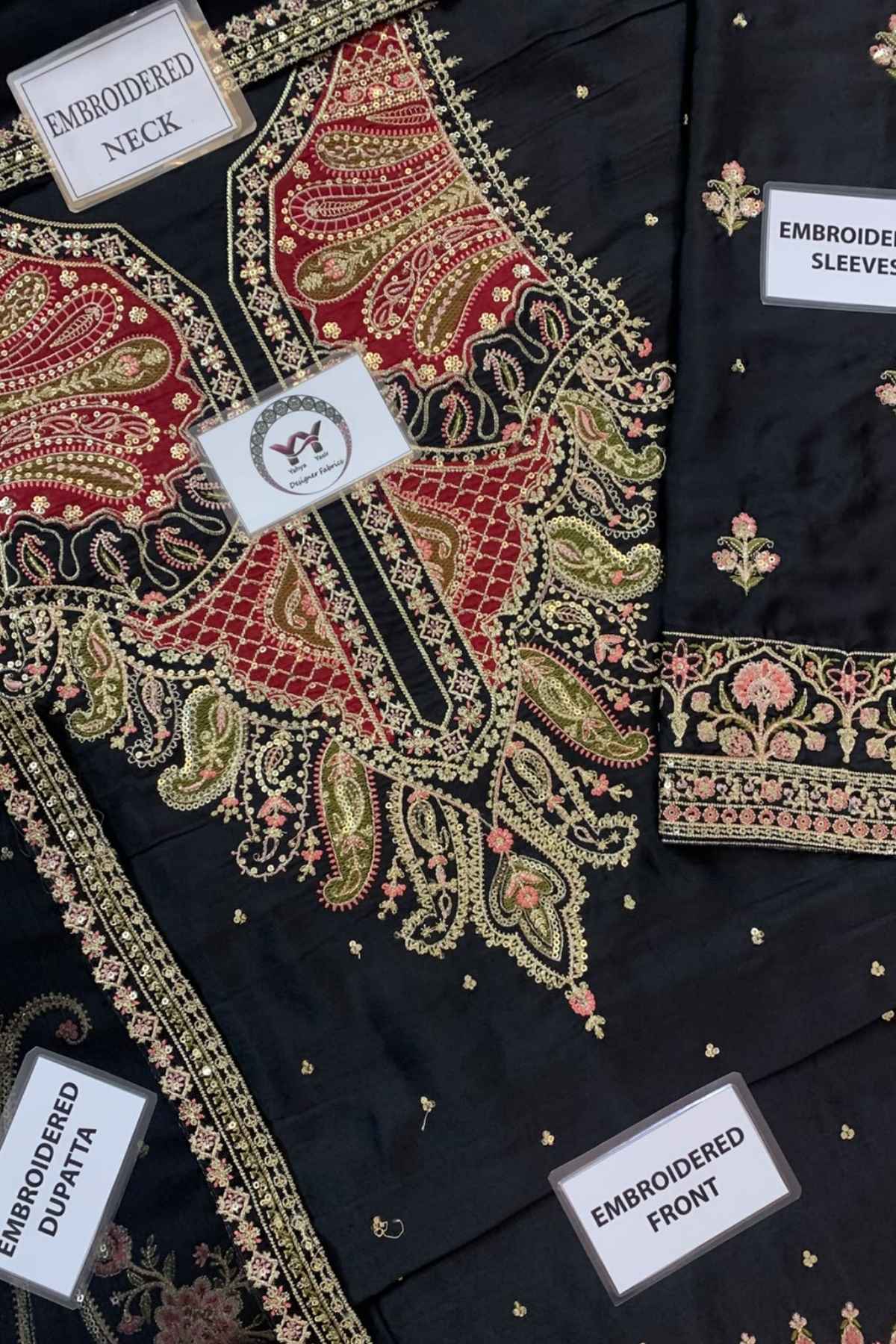 Black embroidered women's salwar suit fabric set with detailed red, gold, and pink paisley and floral embroidery. The image shows close-up views of the neckline, sleeves, front panel, and dupatta, each labeled with a white tag.