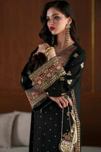 A woman models a black traditional outfit with ornate gold and red embroidery, holding a matching embellished handbag in an indoor setting with a wooden wall background.