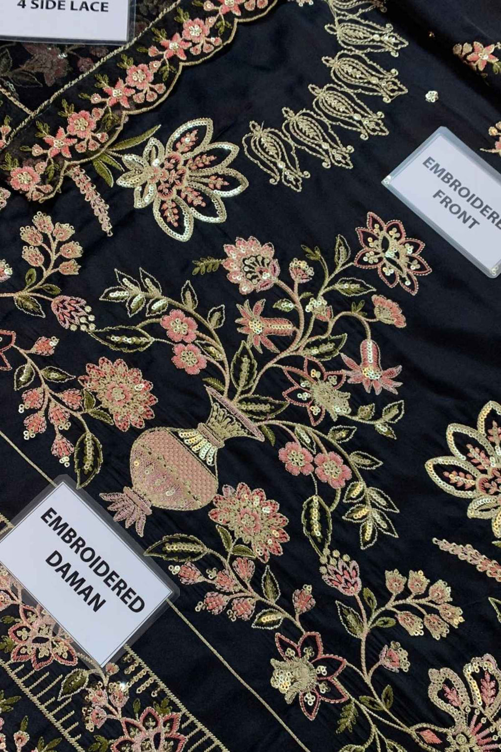 Black embroidered kurta fabric laid flat, showing detailed floral embroidery in pink, gold, and green, with multiple white labels pointing to different embroidered sections such as the neckline, front, and cup.