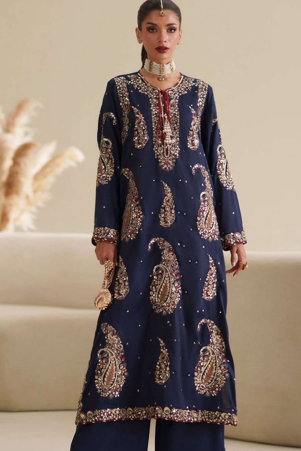 A woman models a navy blue traditional kurta set with intricate gold and silver paisley embroidery, paired with matching navy wide-leg trousers, in a warm beige indoor setting.