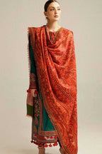 A woman modeling a traditional South Asian outfit featuring a vibrant orange-red patterned dupatta draped over a deep teal green kurta with ornate red and gold borders, set against a plain light gray studio background.