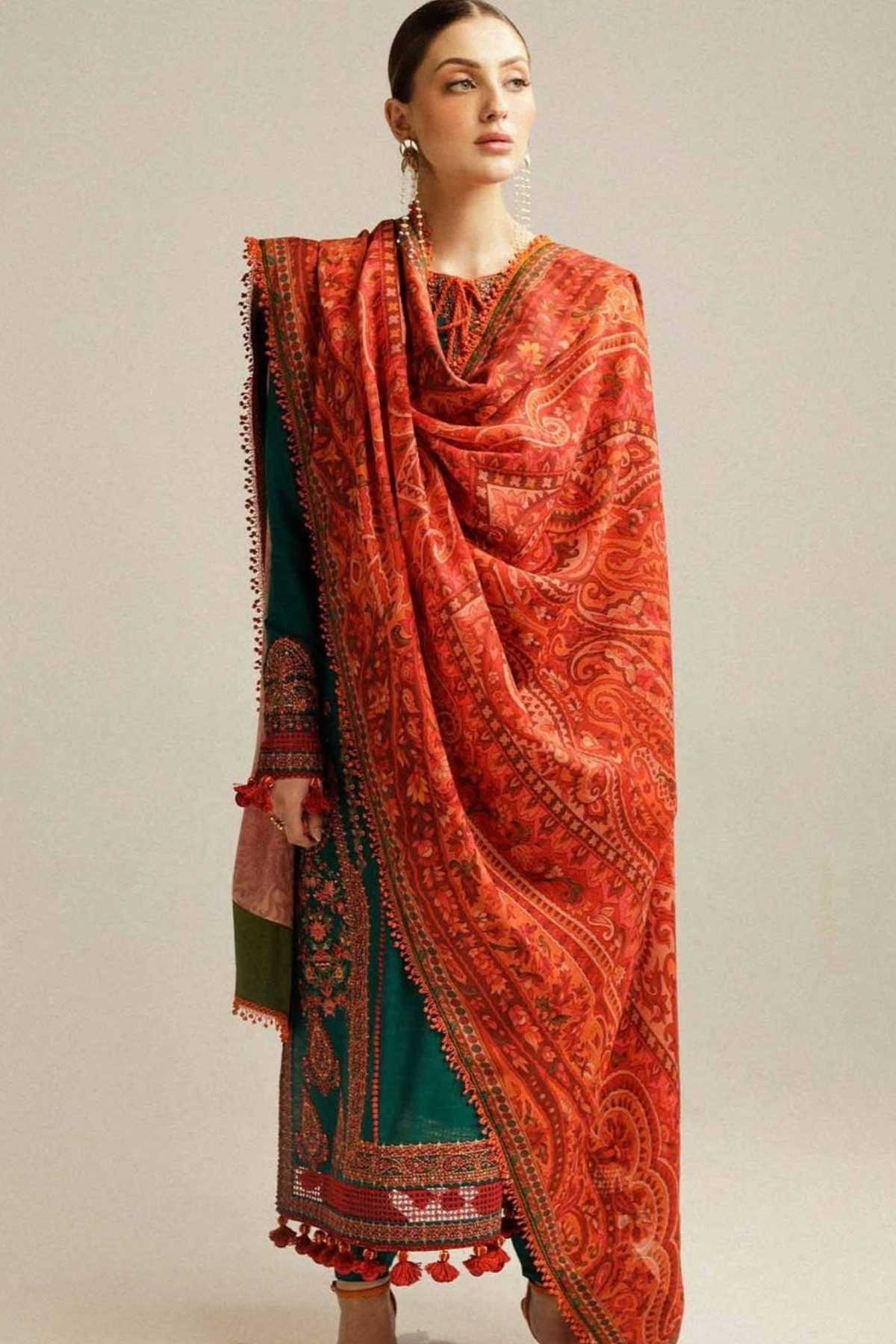 A woman modeling a traditional South Asian outfit featuring a vibrant orange-red patterned dupatta draped over a deep teal green kurta with ornate red and gold borders, set against a plain light gray studio background.