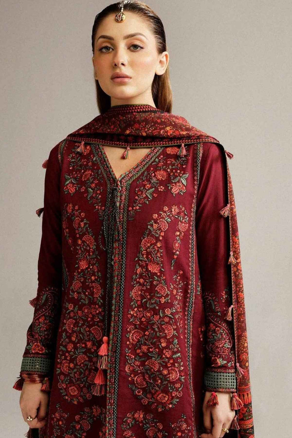 A woman modeling a deep maroon embroidered kurta with a matching embroidered dupatta, featuring intricate floral patterns and tassel details against a neutral gray background.
