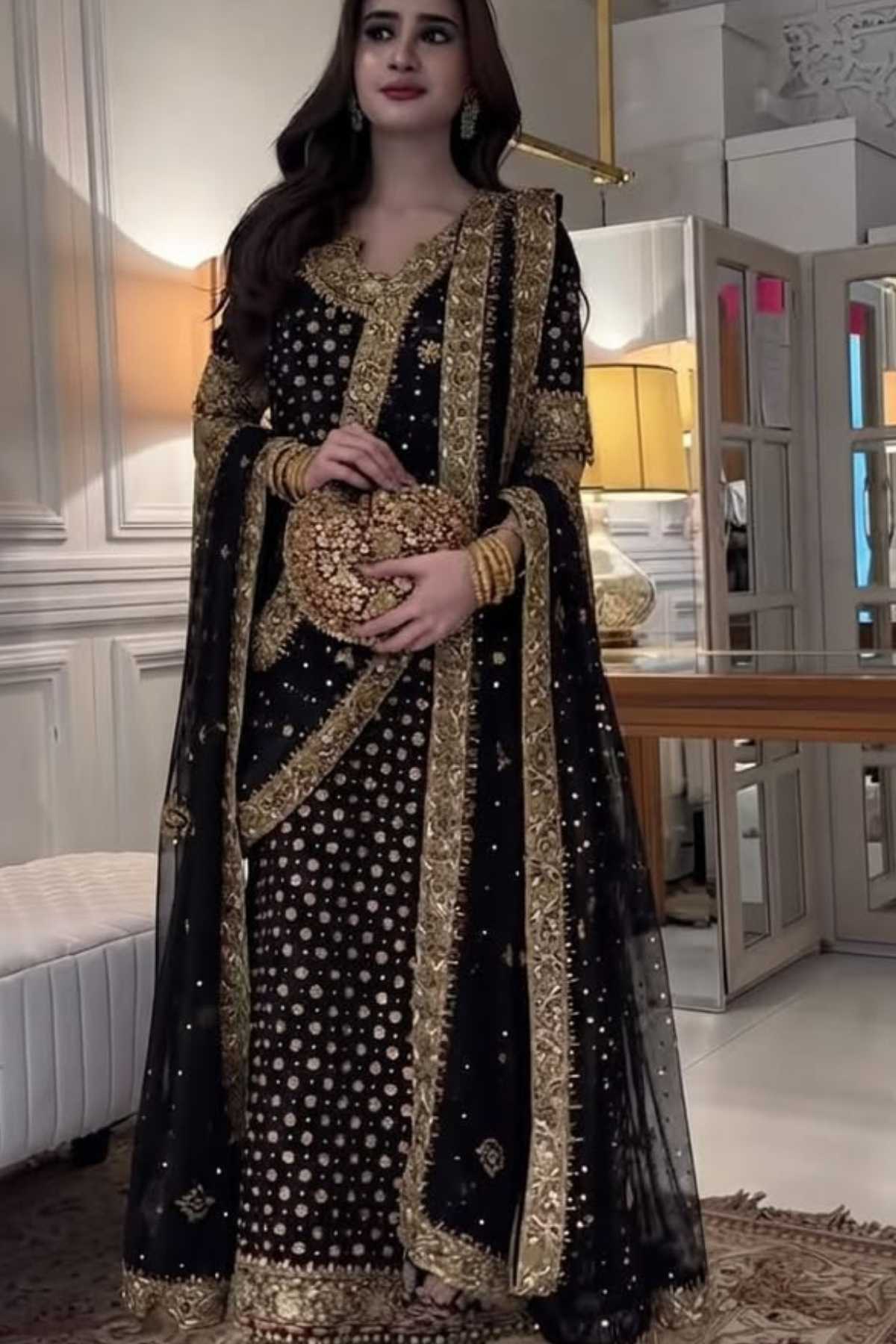 A woman models a black and gold traditional embroidered dress with a matching sheer dupatta, holding a gold clutch in an indoor room setting.
