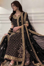 A woman models a black and gold embroidered traditional outfit with a matching sheer dupatta in an indoor setting with white paneled walls.