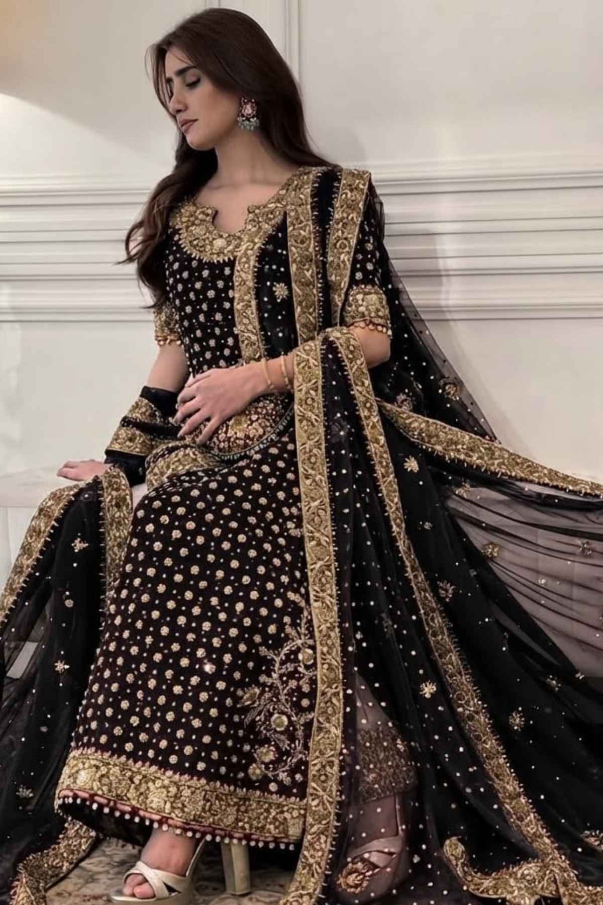 A woman models a black and gold embroidered traditional outfit with a matching sheer dupatta in an indoor setting with white paneled walls.