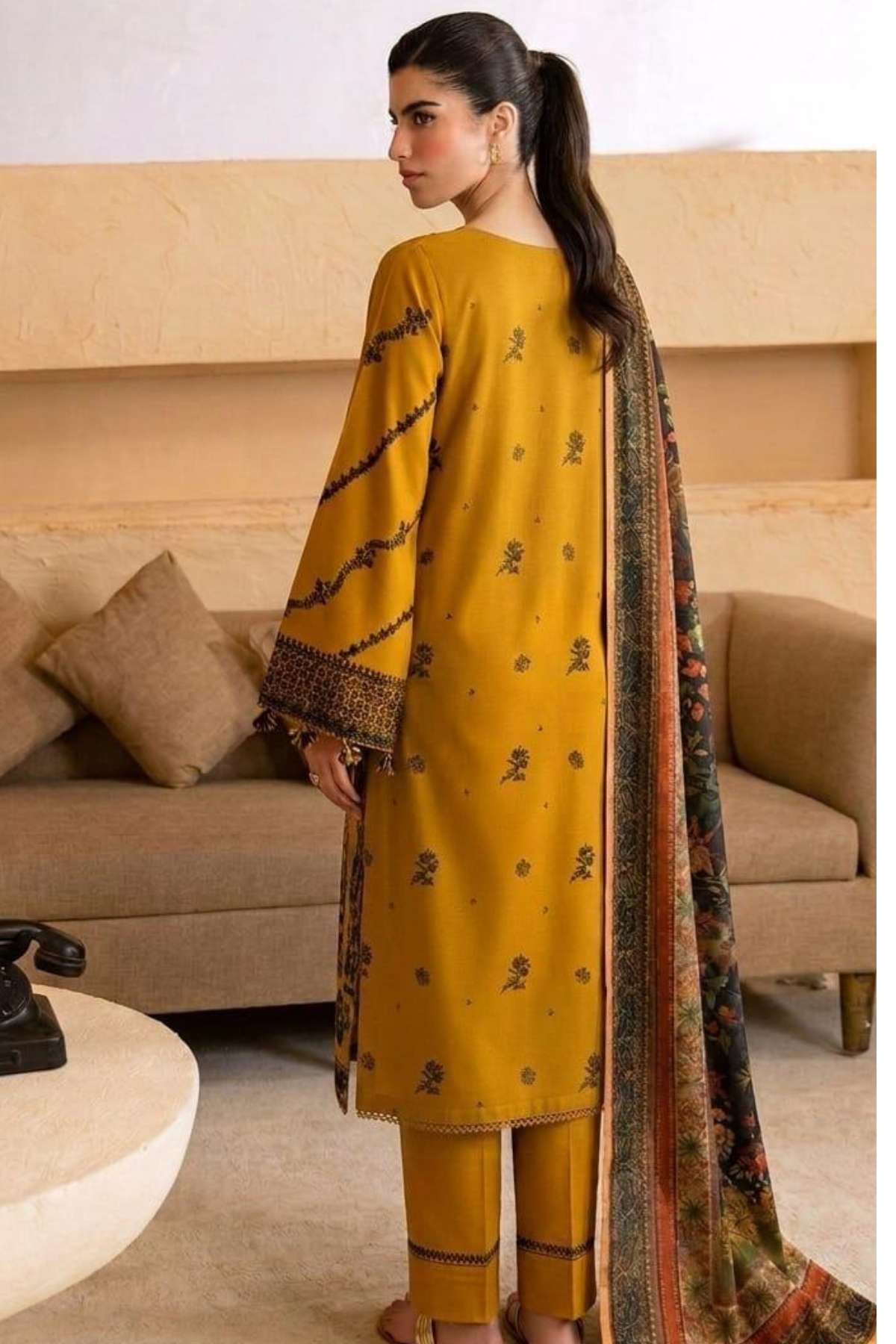 A woman modeling a mustard yellow three-piece salwar kameez suit with black printed motifs and a matching patterned dupatta in a living room setting.