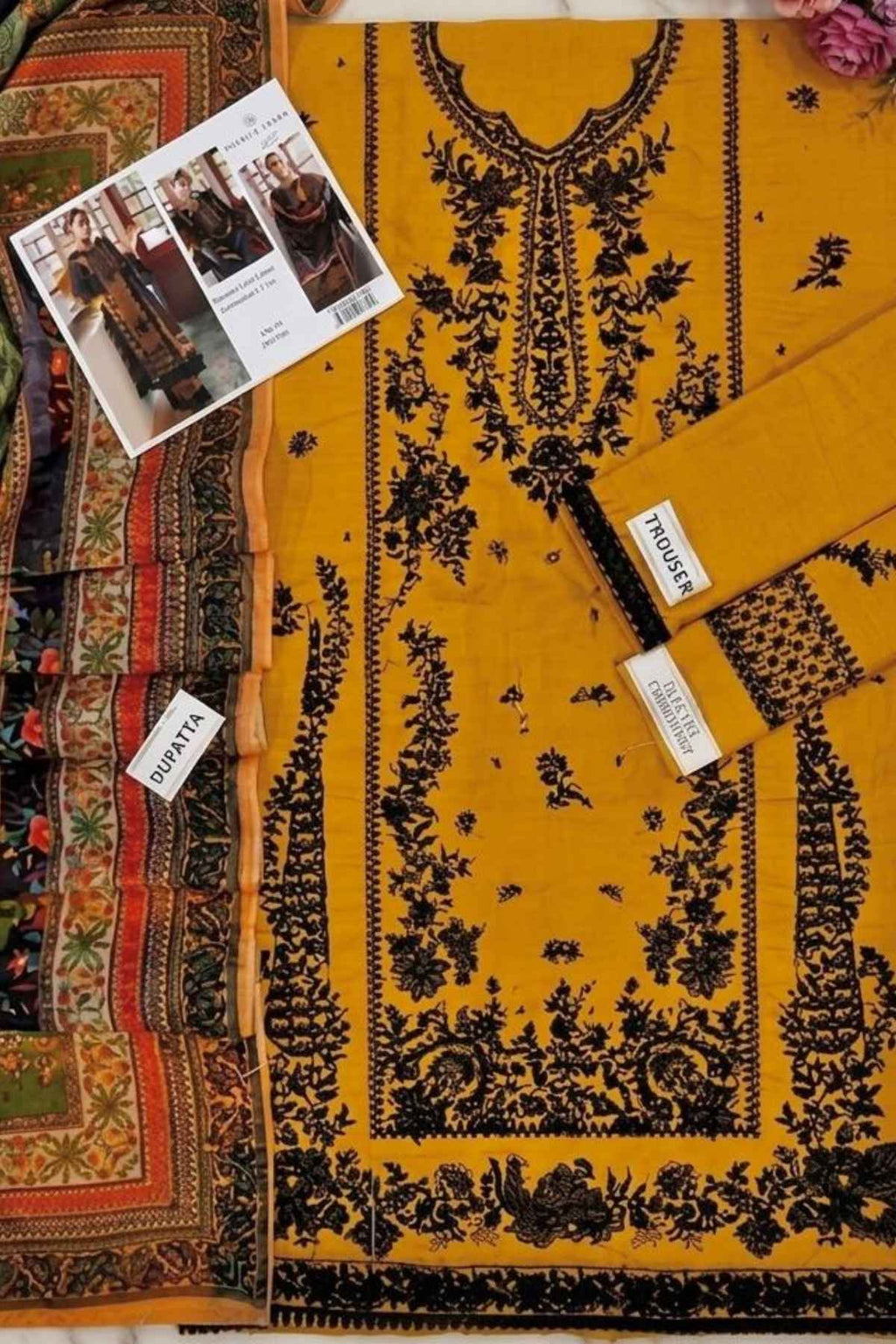 A flat-lay presentation of a yellow unstitched salwar suit set, featuring a heavily embroidered kurta with black floral patterns, matching yellow trousers, a patterned dupatta, and a brand card on a neutral background.