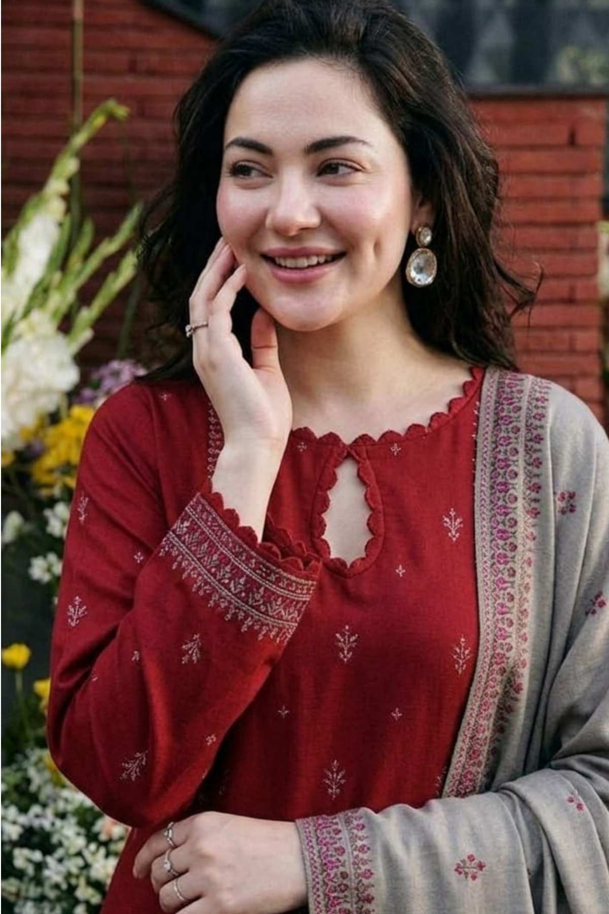 A woman wearing a red embroidered kurta with a keyhole neckline and a beige dupatta, posing outdoors in front of a brick wall and flowers.