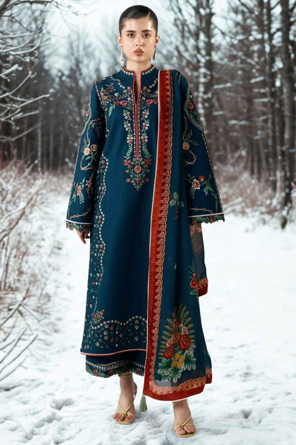 A woman stands in a snowy outdoor setting wearing a long dark teal embroidered kurta with ornate multicolor floral and geometric patterns and a red border panel.