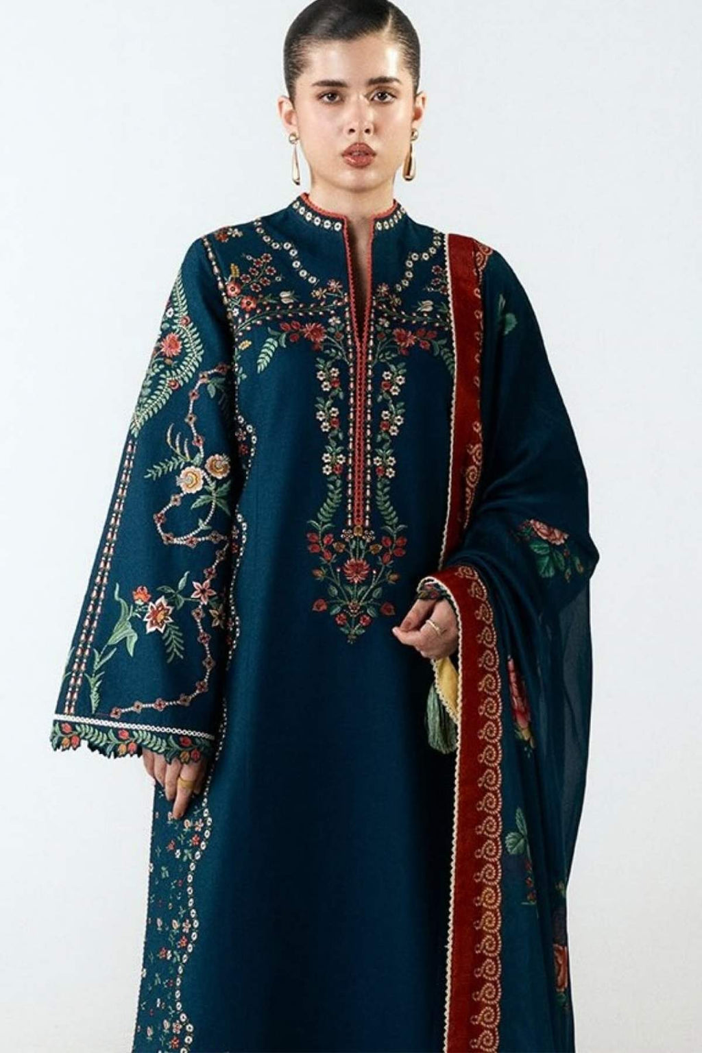 A woman modeling a dark teal traditional embroidered kurta with a matching dupatta. The kurta features intricate floral embroidery in red, gold, and green on the chest, sleeves, and hem, with a red border on the dupatta.