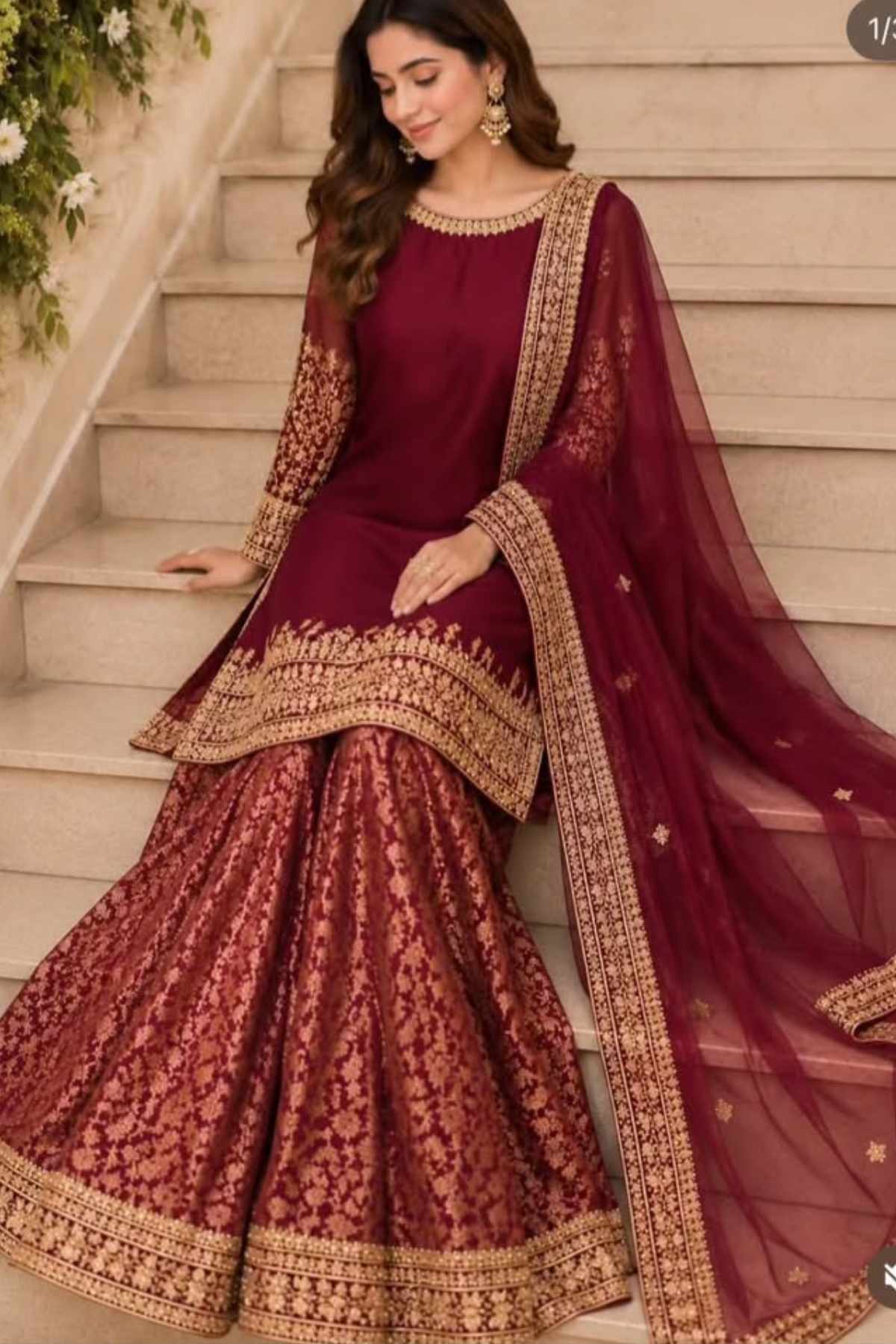 A woman models a traditional maroon and gold embroidered lehenga choli set with a matching dupatta, posed on outdoor stone steps.