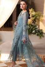 A woman models a traditional South Asian outfit featuring a dusty blue-green long kurta with intricate floral embroidery and a matching sheer dupatta, set against an elegant indoor backdrop with draped curtains and floral arrangements.