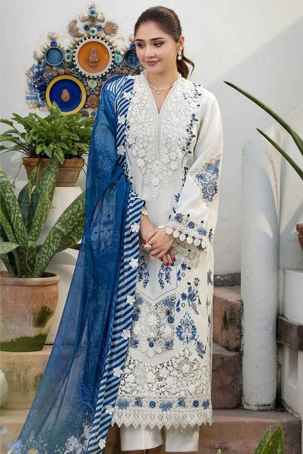 A woman models a traditional South Asian salwar kameez suit. The outfit consists of an off-white embroidered kurta with blue floral patterns and white tassel trim, paired with matching off-white trousers and a blue striped dupatta.