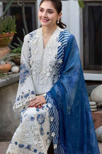 A woman models a traditional South Asian outfit featuring a white embroidered kurta with blue floral patterns, a matching white embroidered skirt, and a sheer blue dupatta with blue stripes and floral motifs, set against an outdoor patio background.