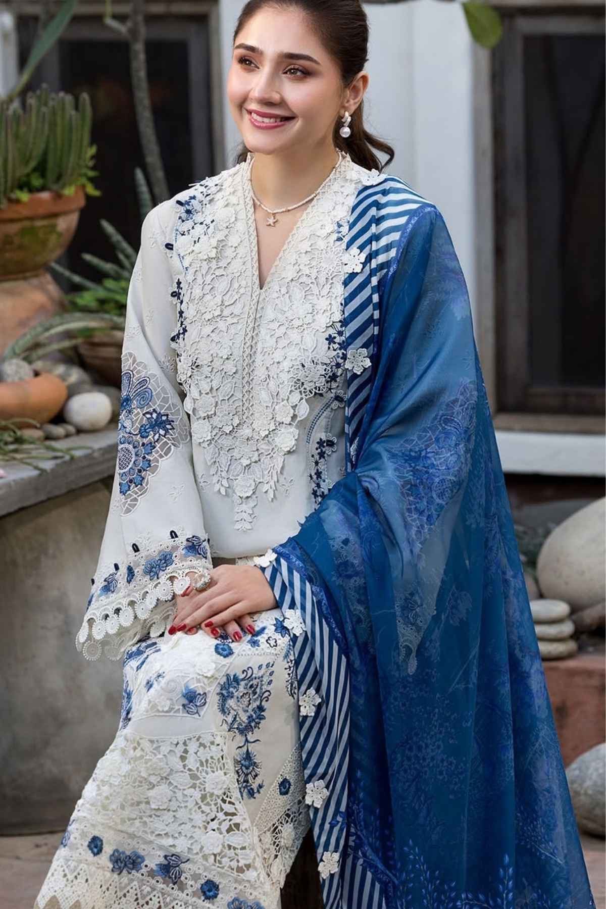 A woman models a traditional South Asian outfit featuring a white embroidered kurta with blue floral patterns, a matching white embroidered skirt, and a sheer blue dupatta with blue stripes and floral motifs, set against an outdoor patio background.