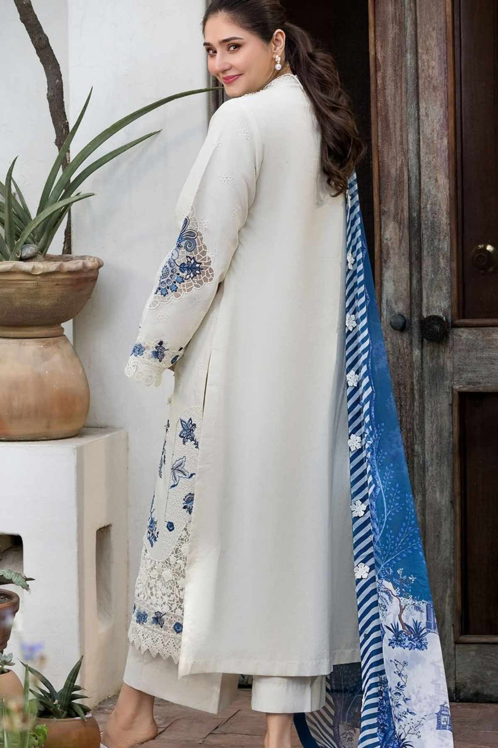 A woman modeling a traditional South Asian outfit featuring a long cream-colored kurta with blue floral embroidery on the sleeves and hem, paired with matching trousers and a blue patterned dupatta, set against a rustic wooden door and potted plants.
