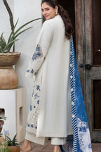 A woman modeling a traditional South Asian outfit featuring a long cream-colored kurta with blue floral embroidery on the sleeves and hem, paired with matching trousers and a blue patterned dupatta, set against a rustic wooden door and potted plants.