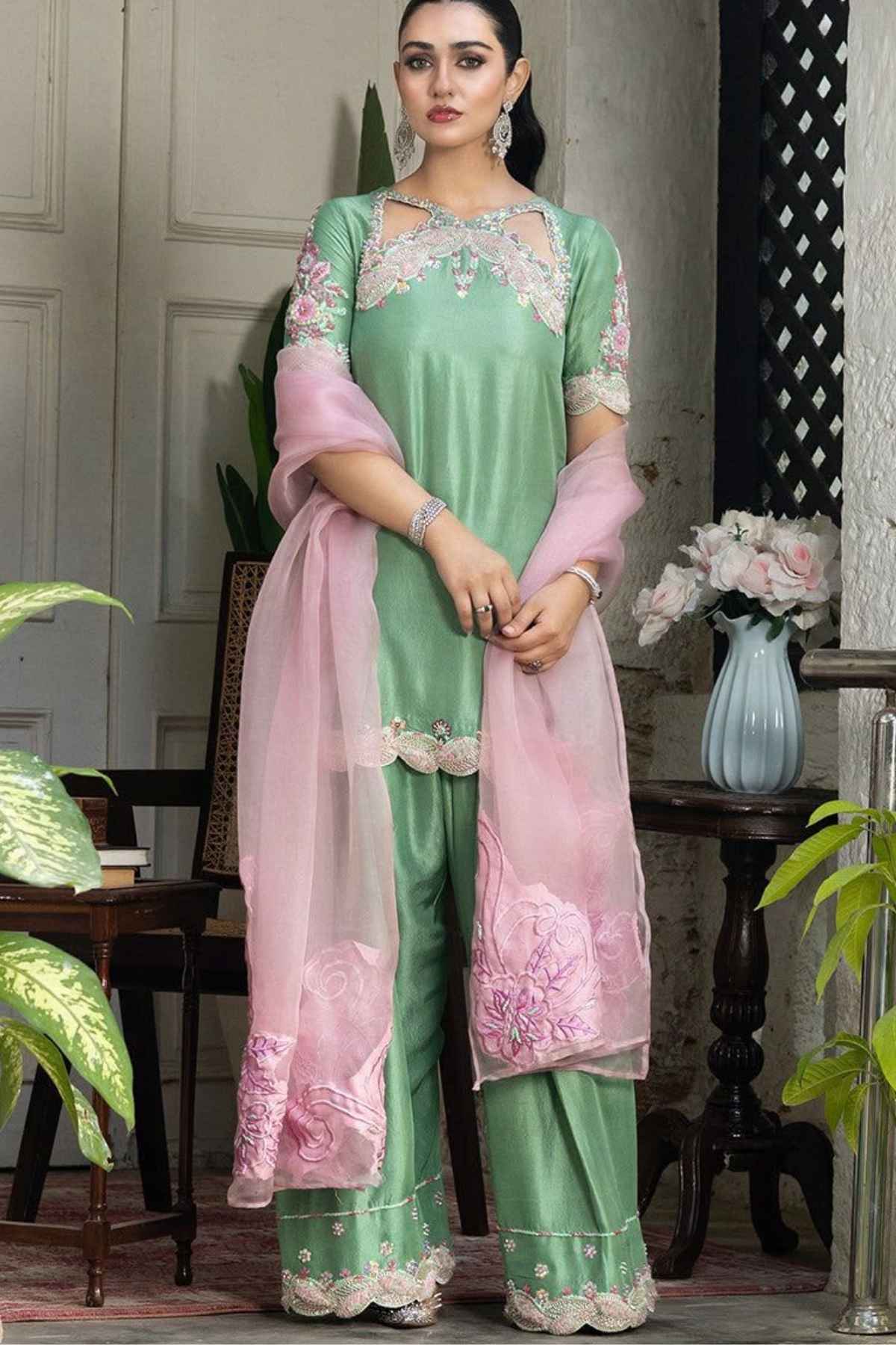 A woman models a mint green and light pink ethnic salwar kameez suit with floral embroidery and lace details, set against an indoor background with plants and furniture.