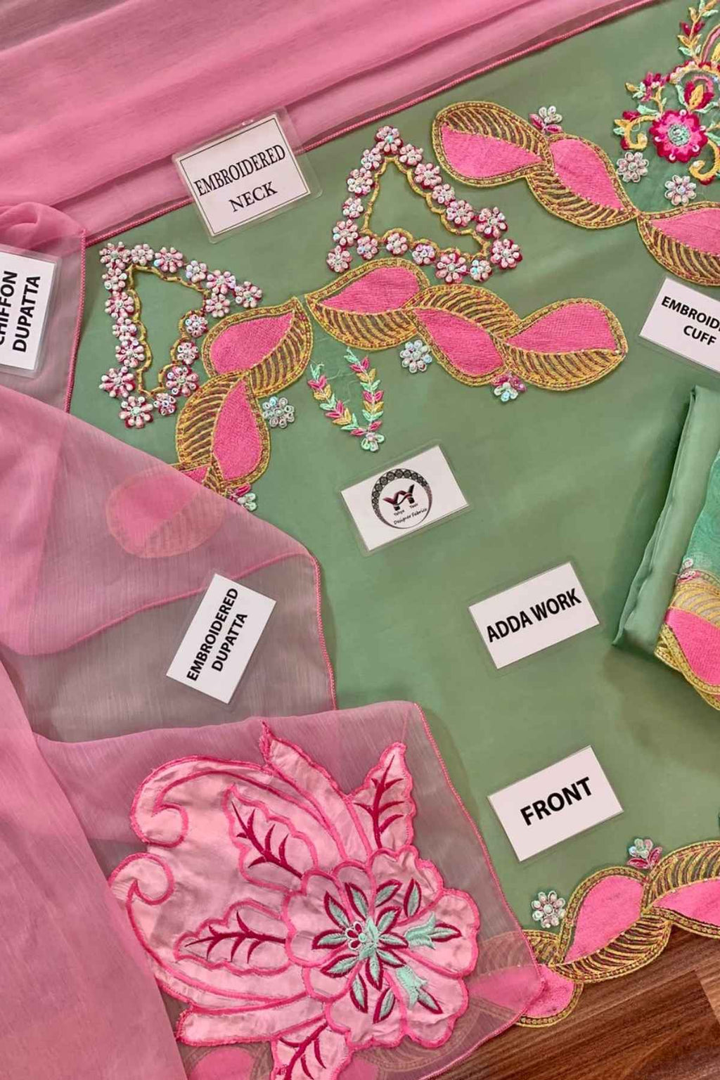 A close-up of a green embroidered fabric garment or textile set with pink and gold decorative embroidery, pink organza dupatta, and several white labels pointing to parts such as embroidered neck, adda work, front, and embroidered dupatta.