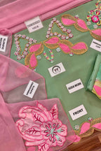 A close-up of a green embroidered fabric garment or textile set with pink and gold decorative embroidery, pink organza dupatta, and several white labels pointing to parts such as embroidered neck, adda work, front, and embroidered dupatta.