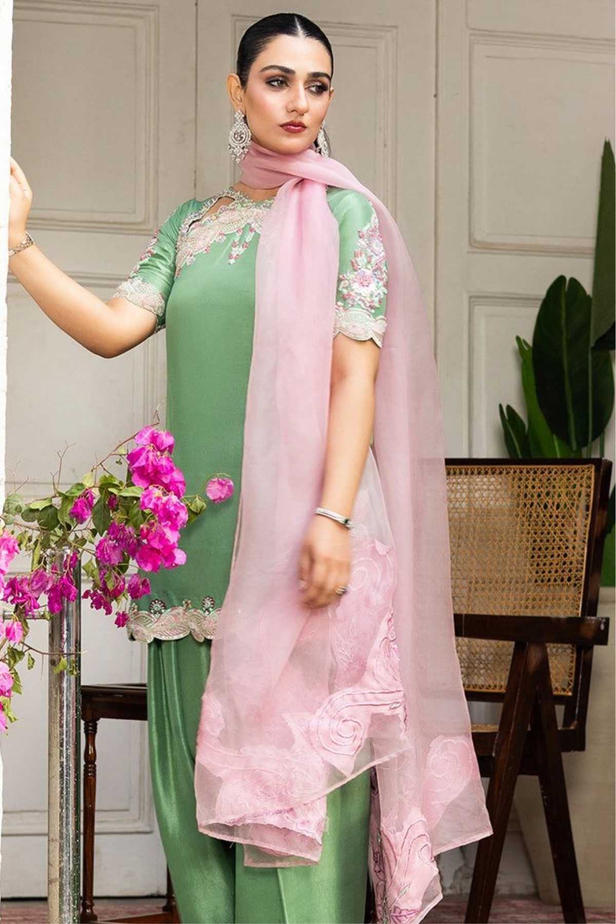 A woman models a traditional Indian salwar kameez suit in mint green and light pink. The outfit features a green kurta with floral embroidery on the sleeves and chest, matching green trousers, and a sheer light pink dupatta draped over her shoulder.