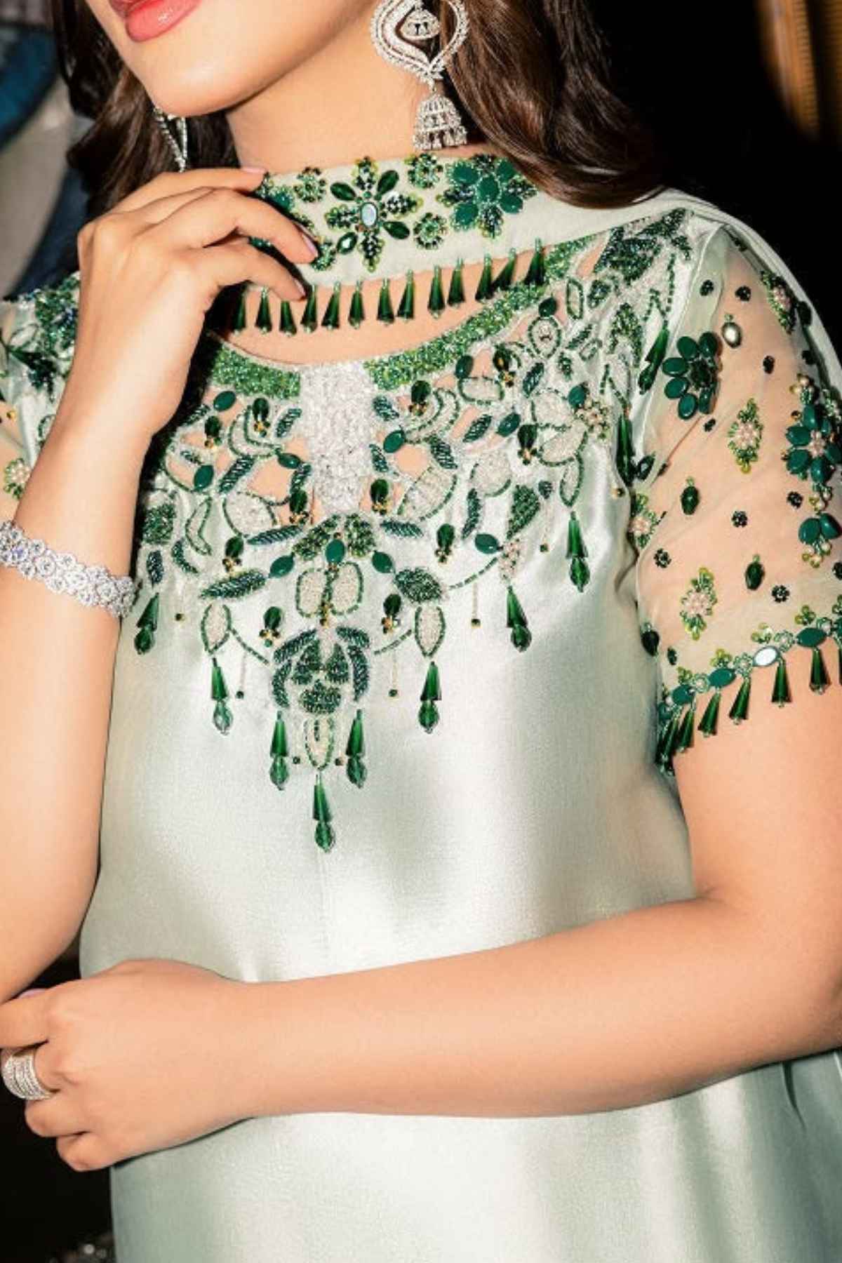 A close-up of a woman wearing a light sage green embellished dress with intricate green and silver embroidery, sheer sleeves, and tassel trim.